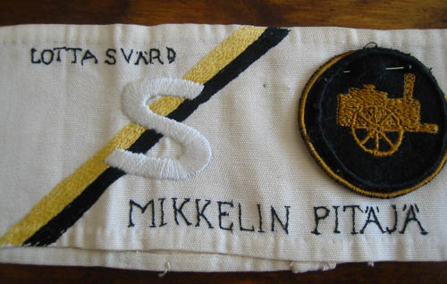 valkoisessa kangasnauhassa keltamusta vinoraita, ommeltuina s-merkki ja kenttkeittimen kuva sek kirjoitettuna tekstit Lotta Svrd ja Mikkelin pitj