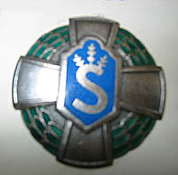 Sinihopeavihre kupera merkki, jossa risti ja havunoksin kruunattu S-kirjain.